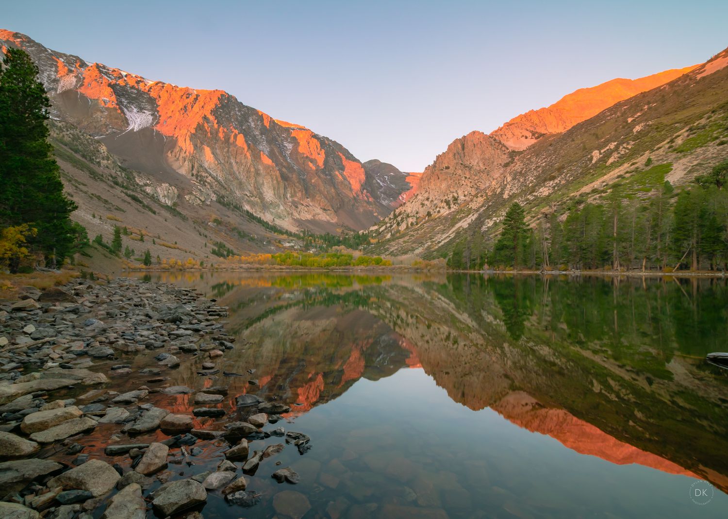 Majestic mountain peaks glow orange at sunrise reflecting in a calm alpine lake surrounded by rocky terrain.
