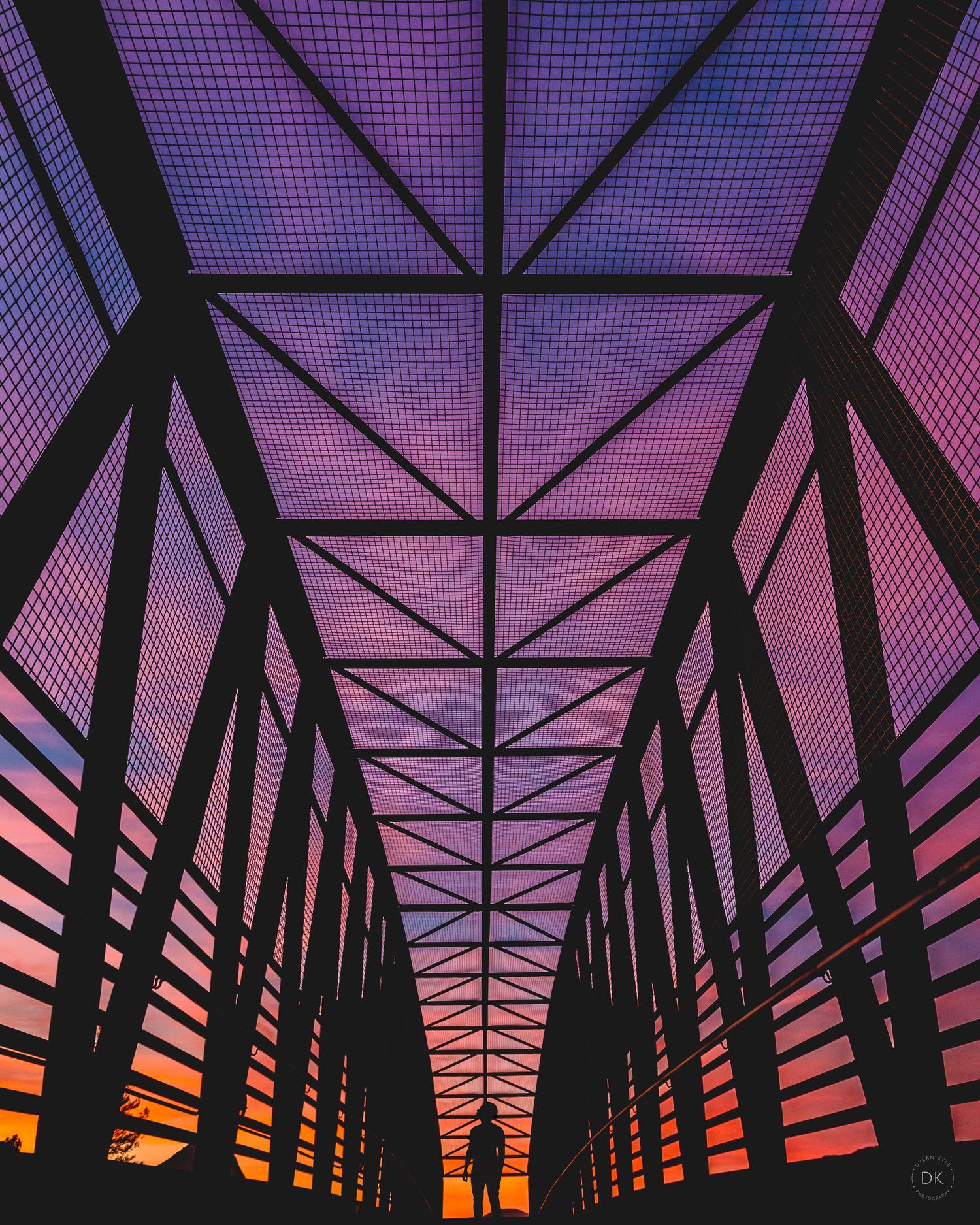 Silhouetted bridge structure against a vibrant purple and orange sunset sky creating a dramatic geometric pattern.