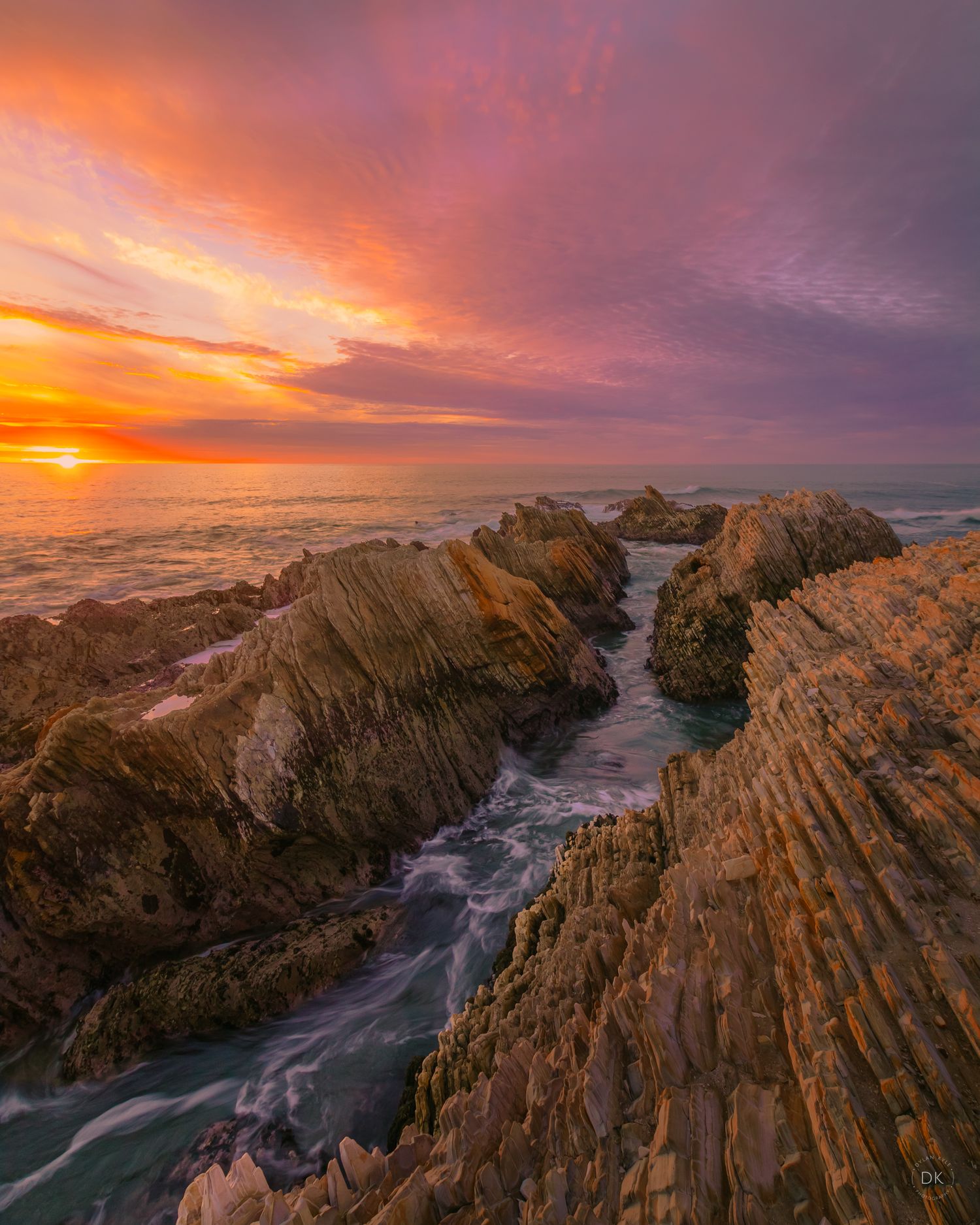 Dramatic sunset over rocky coastline with jagged cliffs and swirling ocean water flowing through rocky channel.