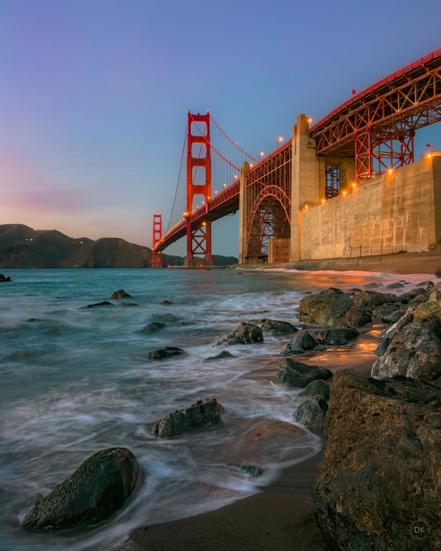 Golden Gate Bridge towers over rocky beach with crashing waves during sunset in San Francisco Bay.