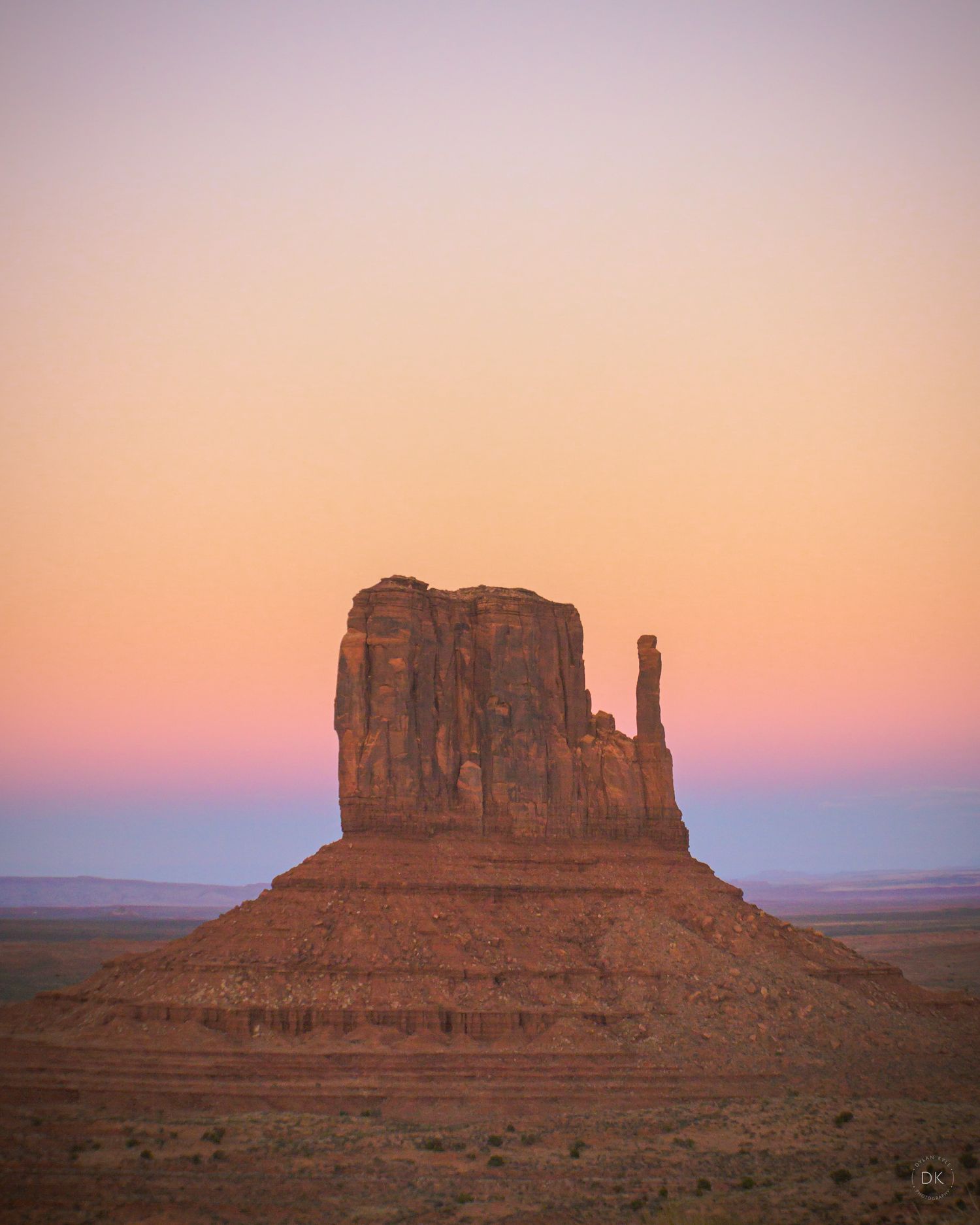 Monument Valley sandstone butte silhouetted against a pastel orange and pink sunset sky in the American Southwest.
