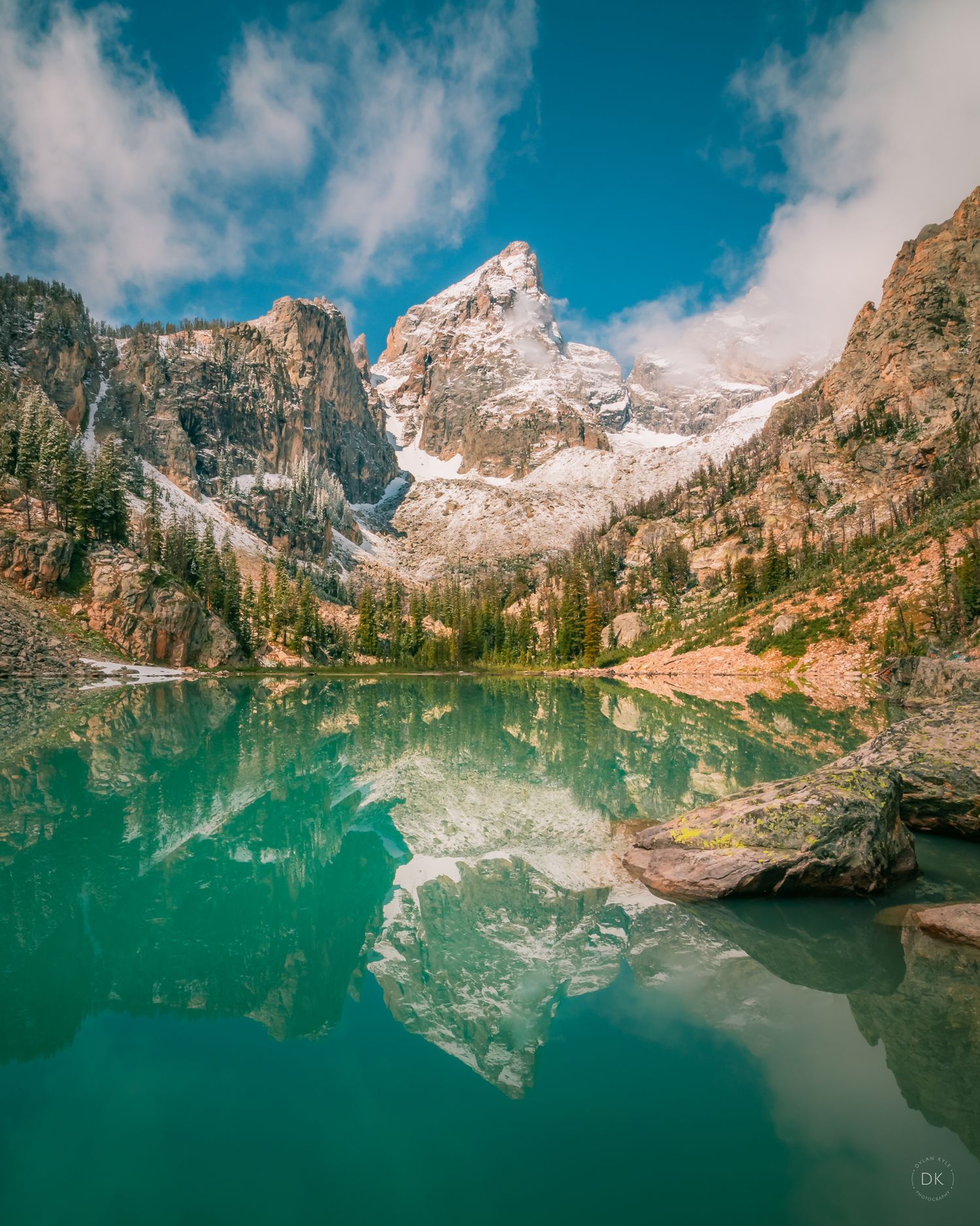 Snow-capped mountain peak reflects in turquoise alpine lake surrounded by evergreen forest and rocky cliffs.