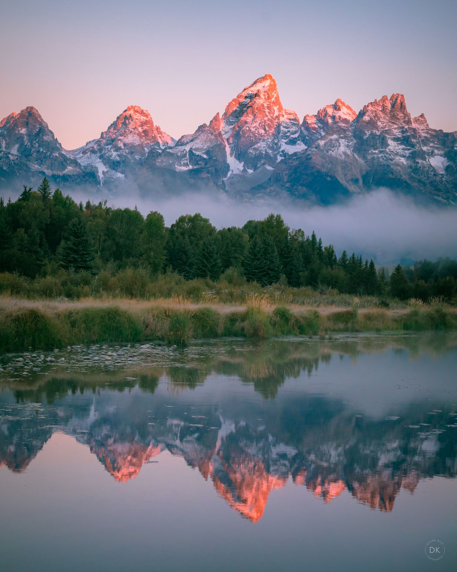 Snow-capped Teton mountains reflect in tranquil lake at sunrise with misty forest and pink-tinged peaks in Wyoming.