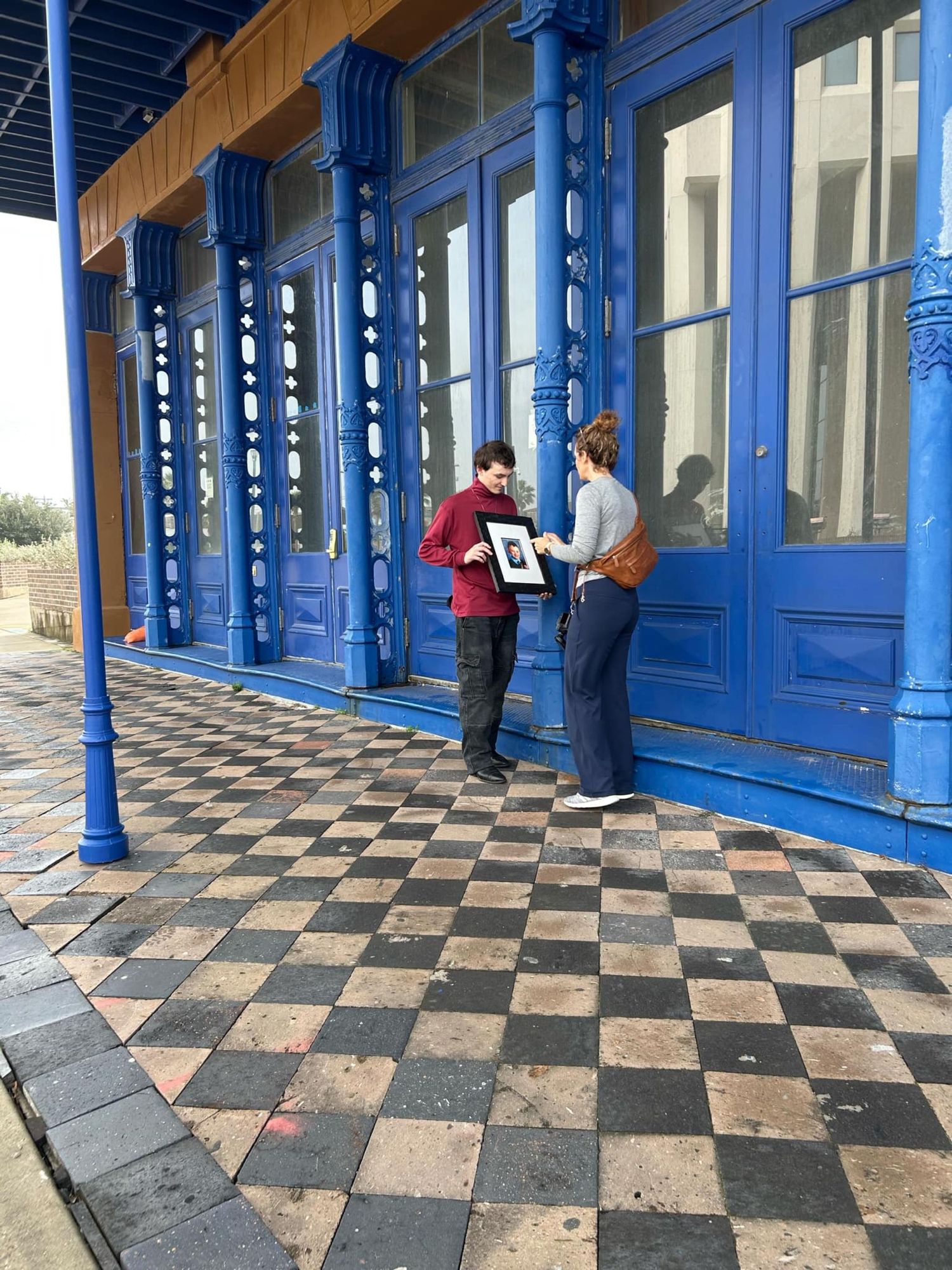 People converse on a historic building's porch with distinctive blue columns and a checkerboard floor pattern.