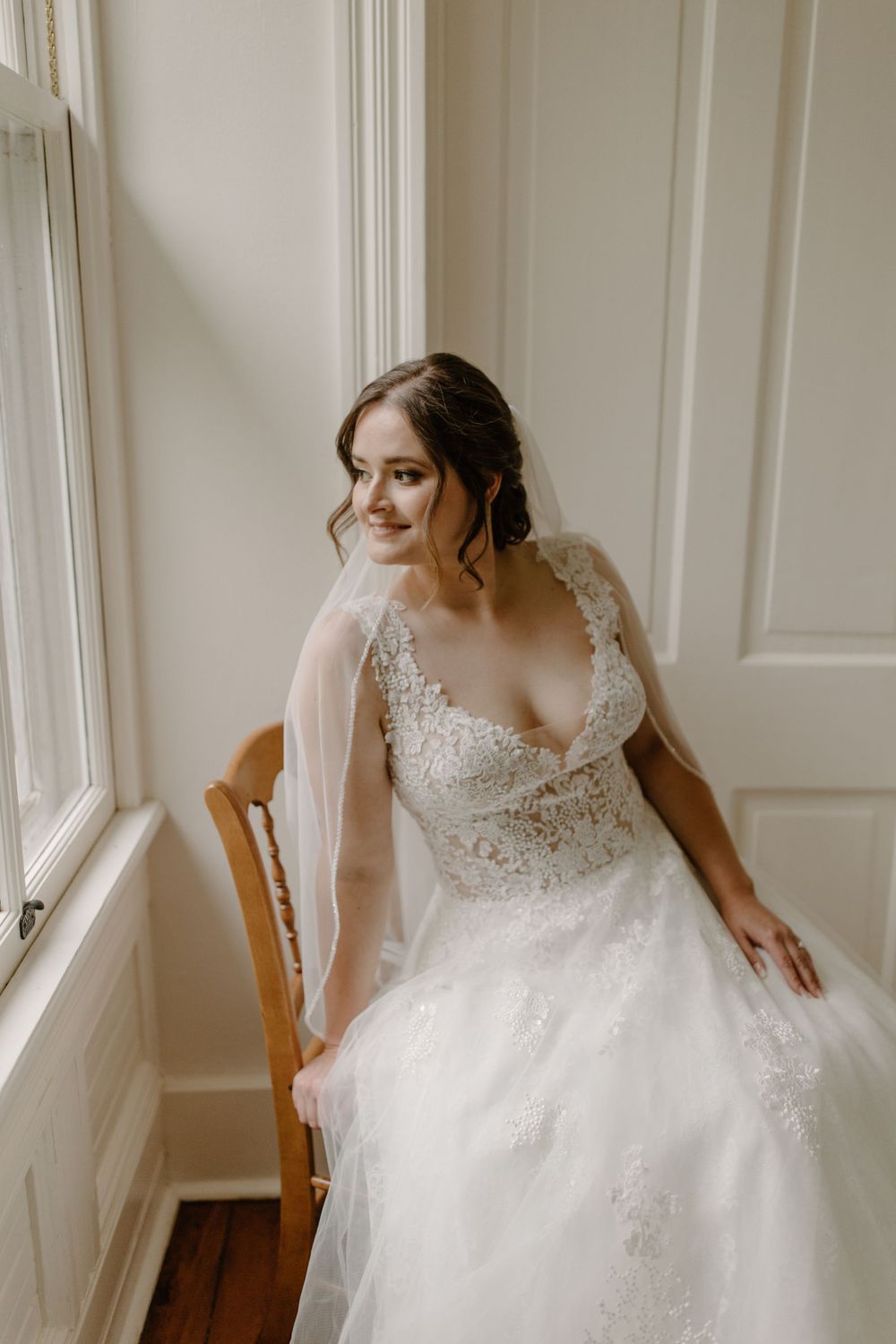 Intimate bridal portrait showing detailed lace wedding dress while seated by a sunny window.