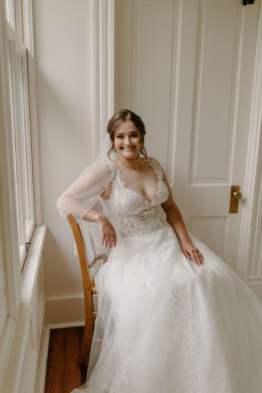 Candid bridal portrait in a white lace wedding gown seated by a bright window.