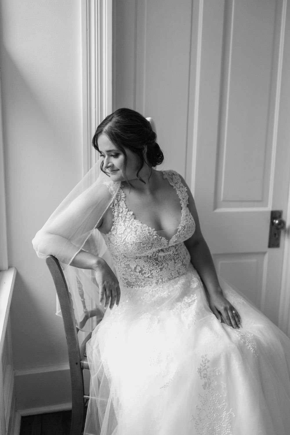 Black and white bridal portrait showing intricate lace details of wedding dress near window.