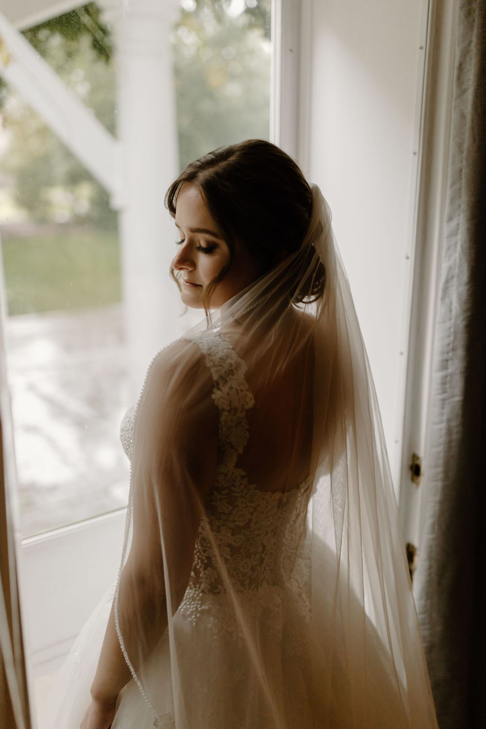 Romantic bridal silhouette by a window featuring an elegant lace wedding dress and long veil.