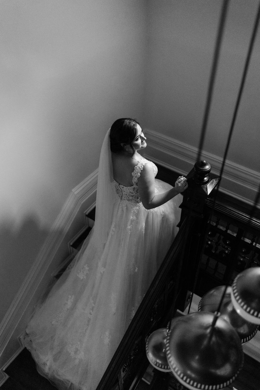 Dramatic black and white shot of bride on staircase with ornate light fixtures below.
