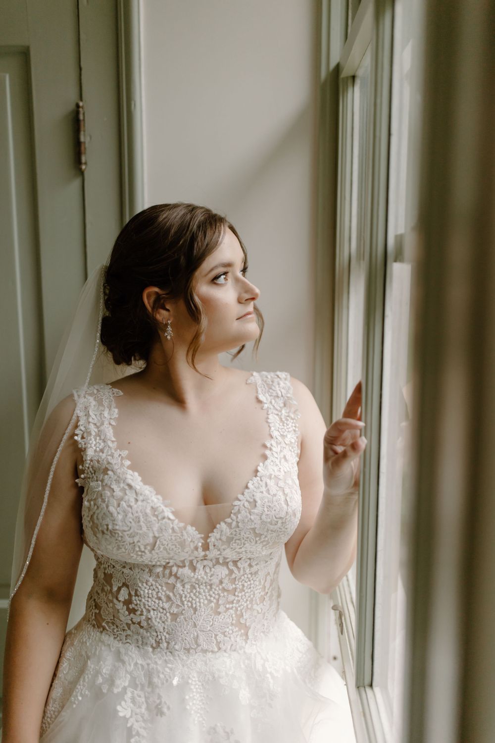 Classic bridal portrait showcasing delicate lace wedding dress with V-neckline by window.