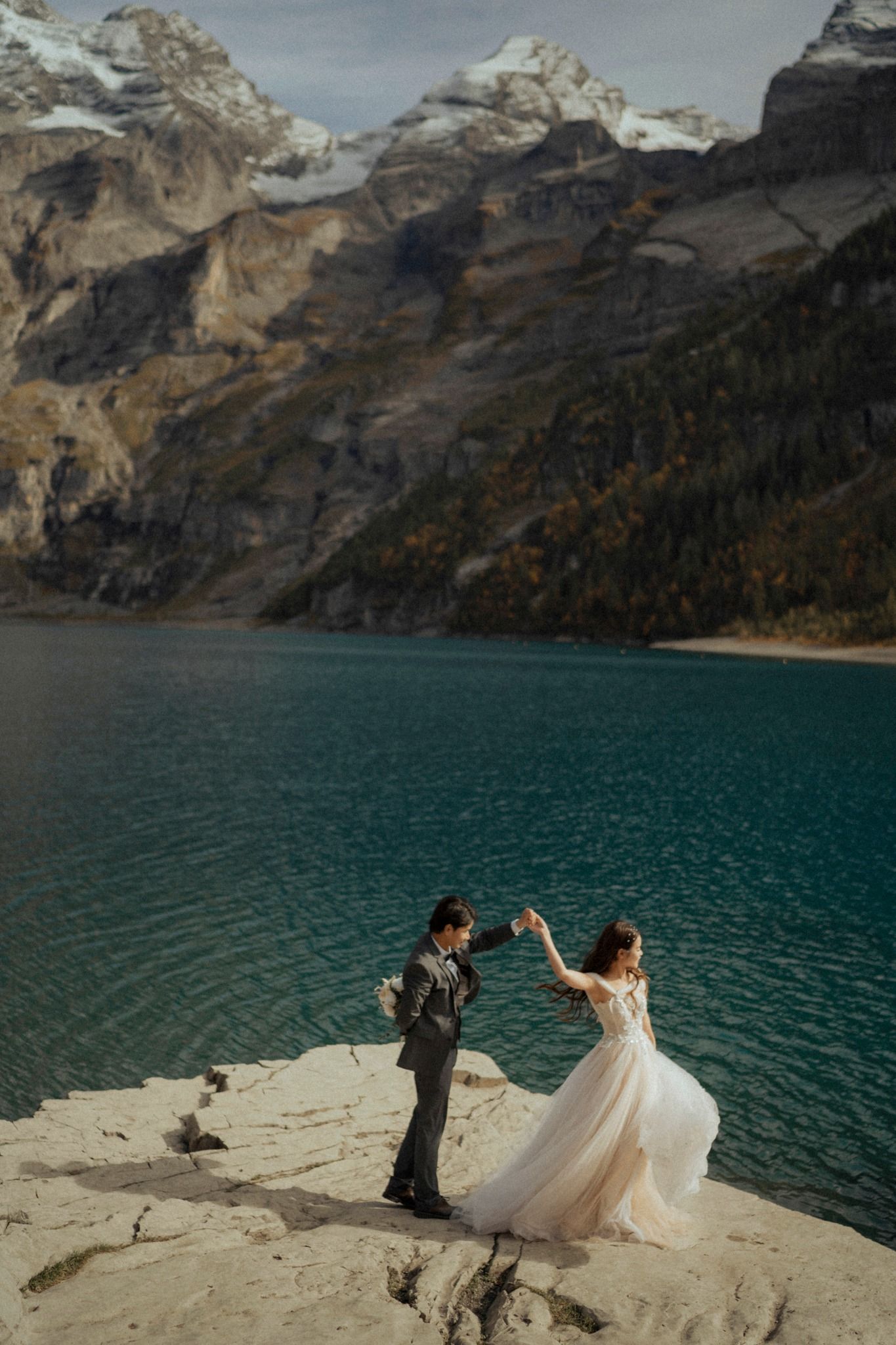 Couple dancing on mountain cliff overlooking turquoise lake and snow-capped peaks.
