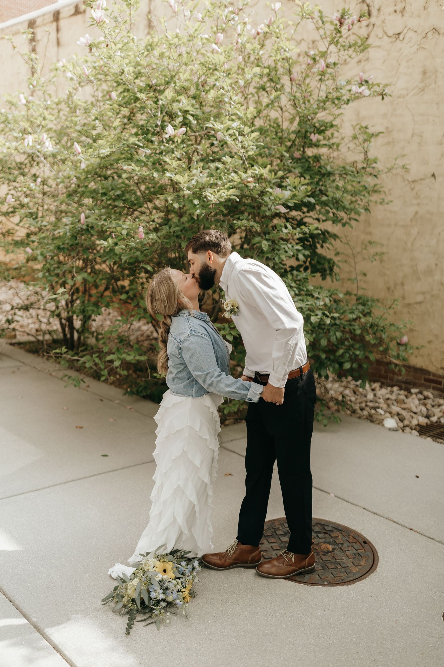 A couple shares a tender kiss near a garden wall during their wedding photoshoot with a rustic bouquet on the ground.
