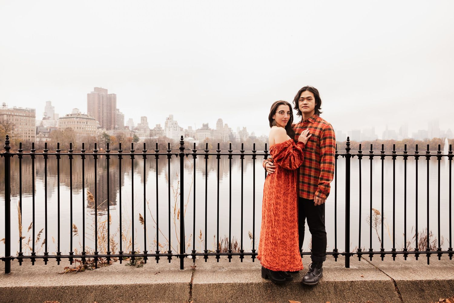 A couple poses along a railing overlooking a misty city skyline.