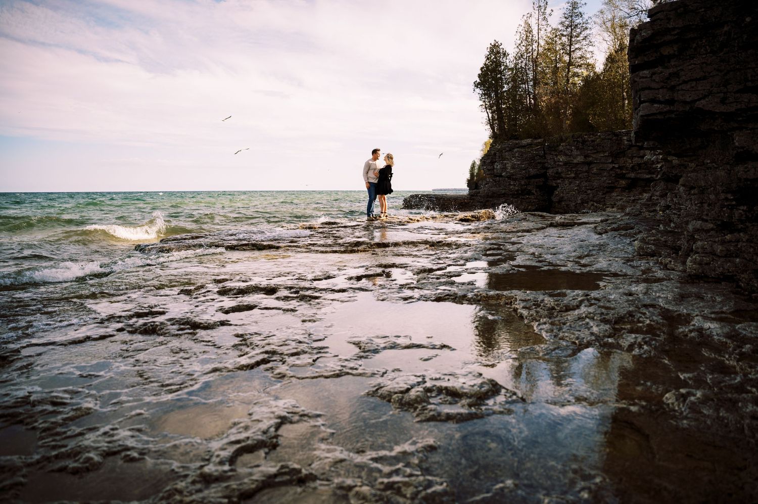 A figure stands on the rocky shoreline of Lake Superior near cliffs and evergreen trees during golden hour.