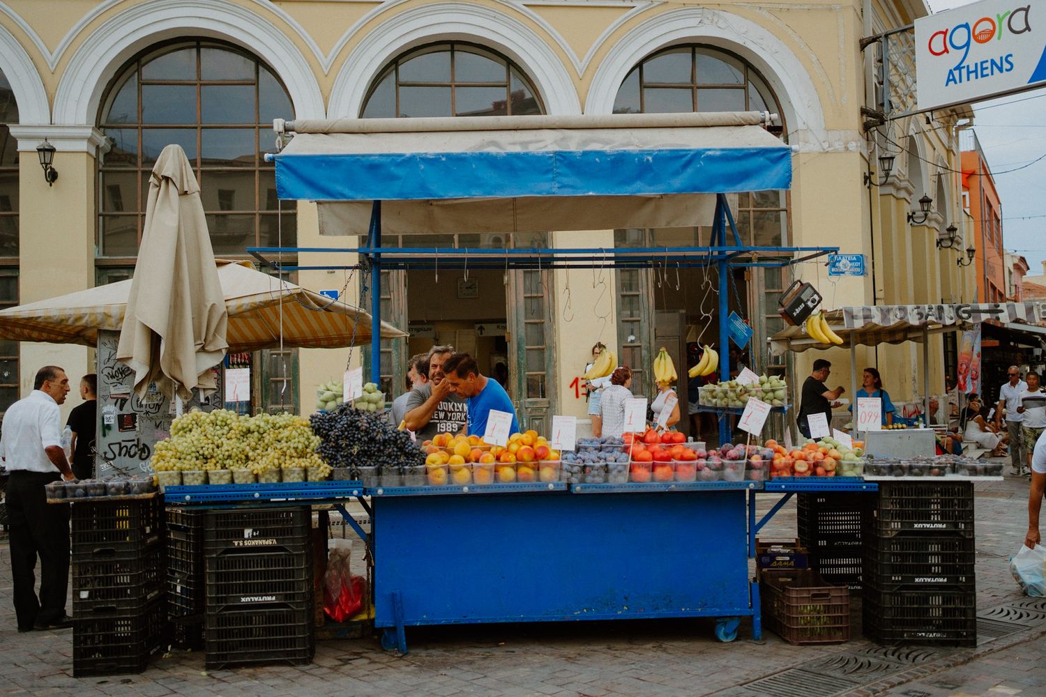 A bright blue fruit and vegetable market stall with colorful produce under historic arched colonnade in a European city.