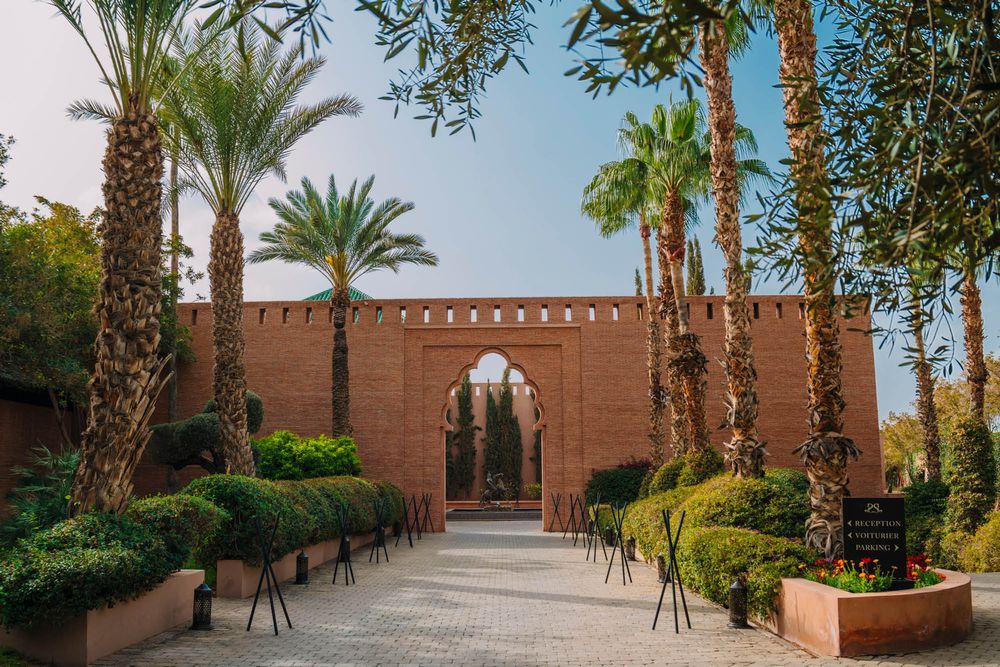 Ornate Moroccan archway entrance with palm trees and manicured gardens along a stone pathway leads to a desert resort.