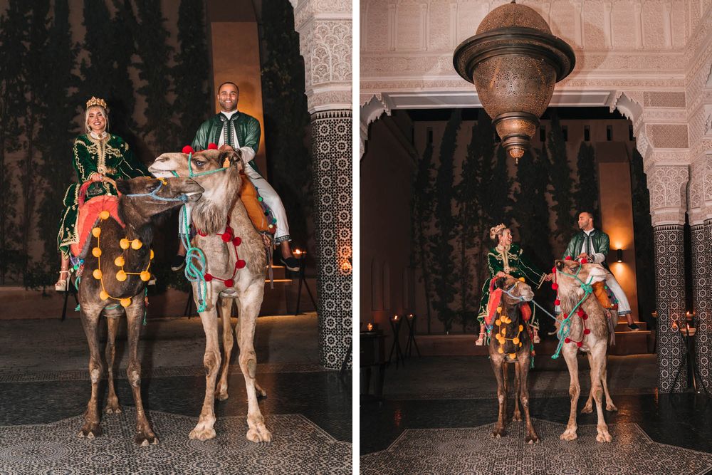 Two people ride decorated camels at night under ornate archways of a historic building.