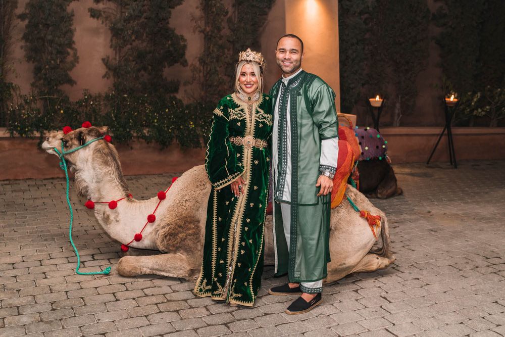 Two people in traditional Moroccan attire pose with a decorated camel on cobblestone ground during evening hours.
