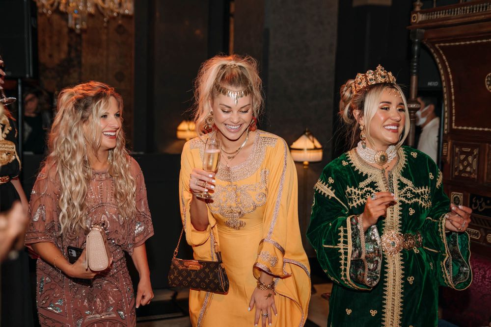 Three friends in colorful boho-style dresses laugh and enjoy drinks at an ornate indoor venue.