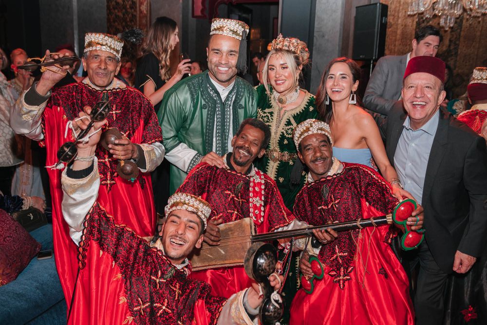 A festive party scene with performers in traditional red and green ceremonial attire celebrating at an indoor event.