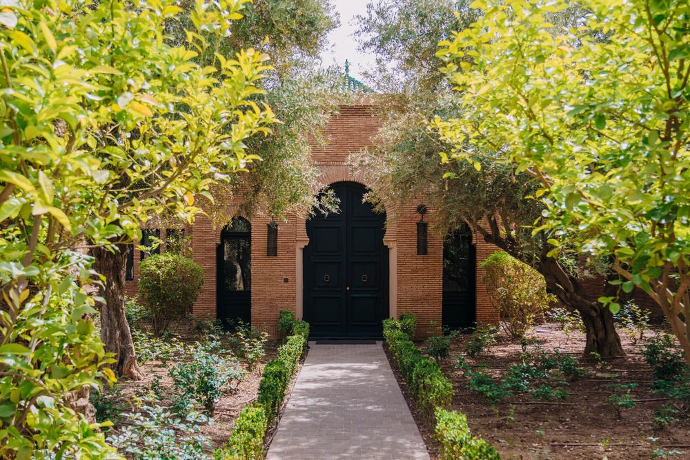 Brick archway entrance with ornate details flanked by lush citrus trees and garden plants along a stone pathway.