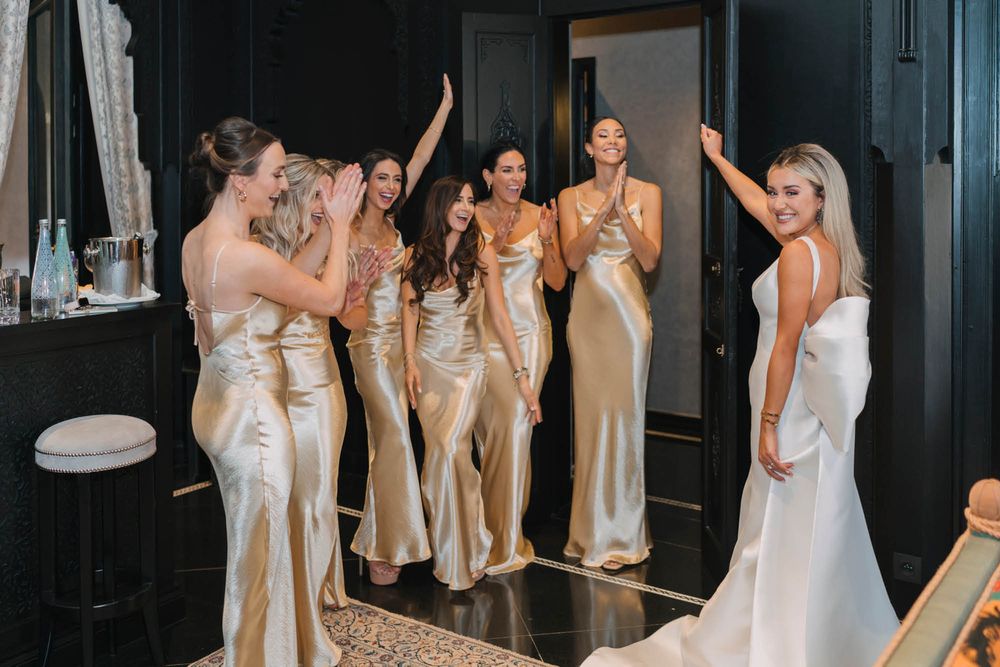 Wedding party in matching champagne satin bridesmaid dresses gather around bride in white gown during pre-ceremony preparations.