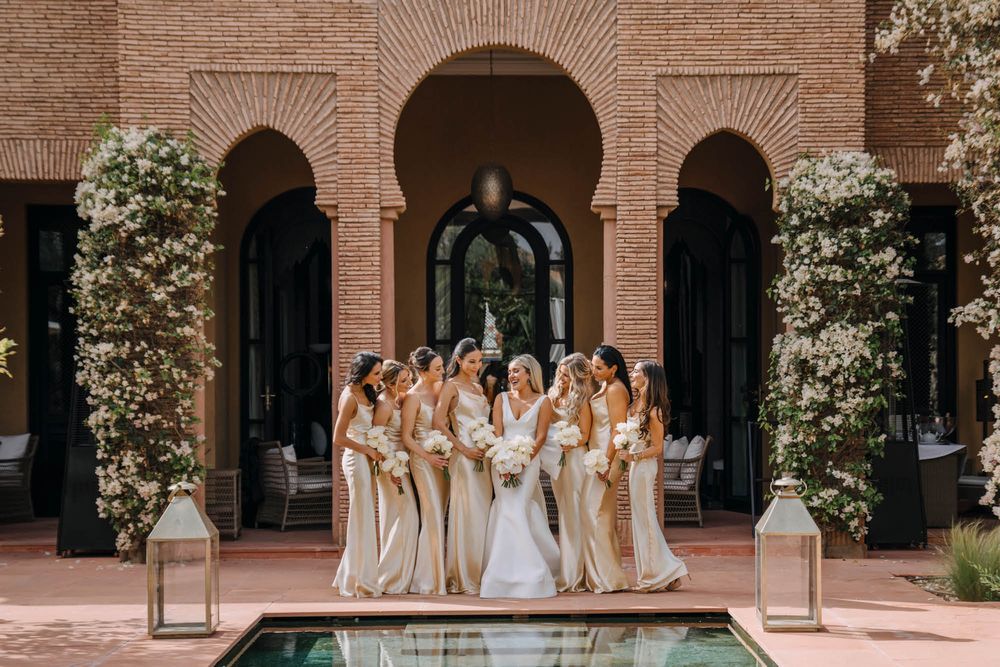 Bridesmaids in champagne dresses gather with bride by elegant archway and reflecting pool at wedding venue.