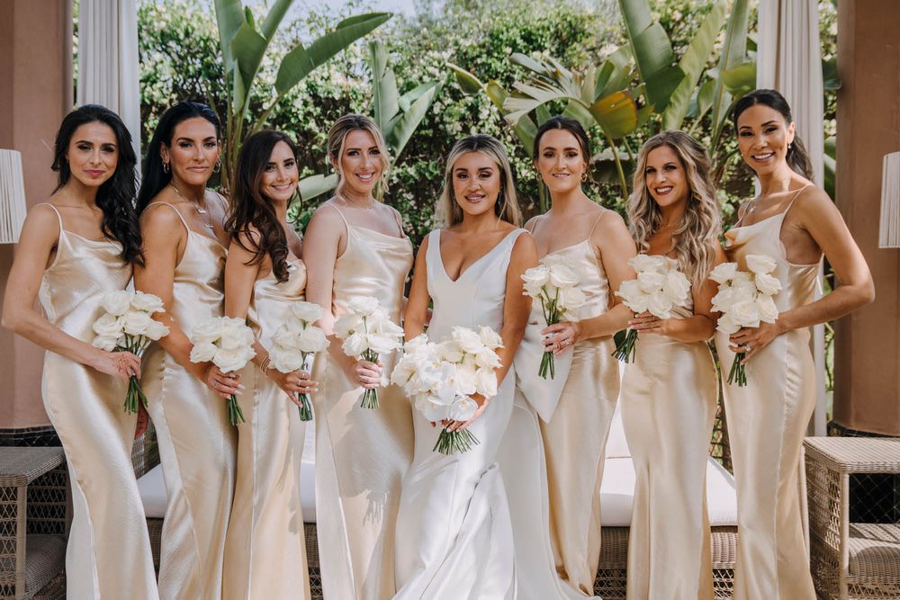 A bridal party in elegant cream-colored silk gowns poses with white bouquets against a tropical garden backdrop.