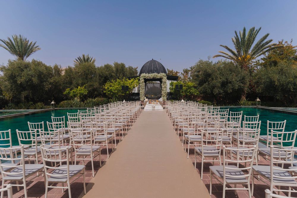 White chairs line a long path leading to an elegant wedding gazebo surrounded by palm trees and gardens.
