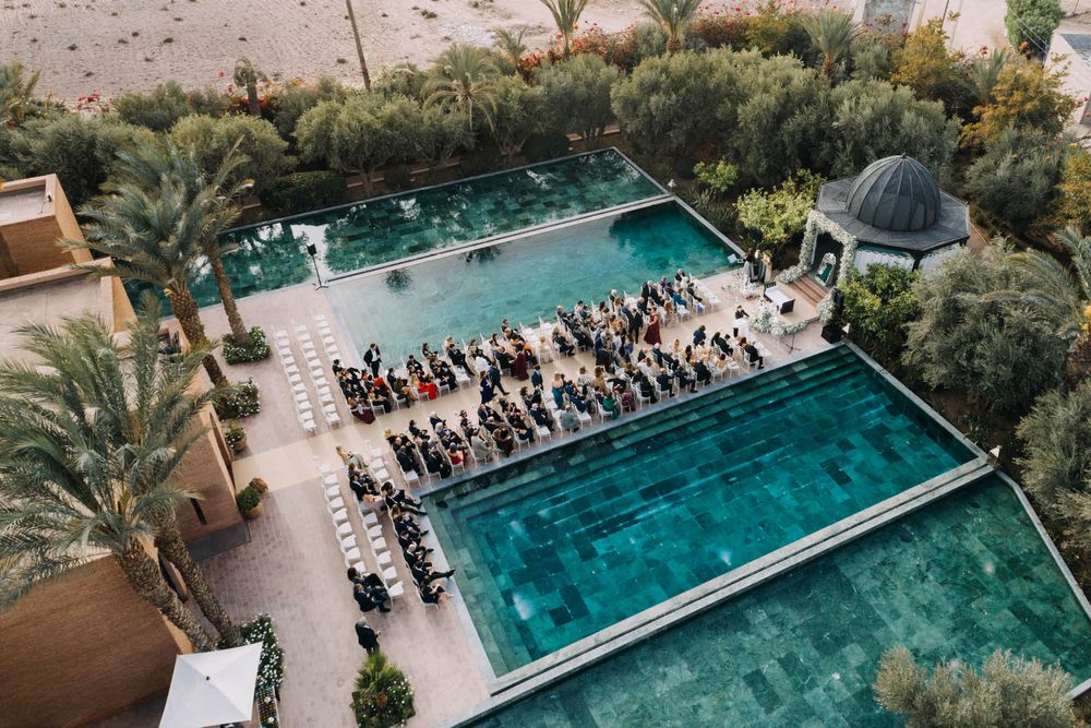 Aerial view of an outdoor wedding ceremony setup between two turquoise pools surrounded by palm trees and desert landscape.