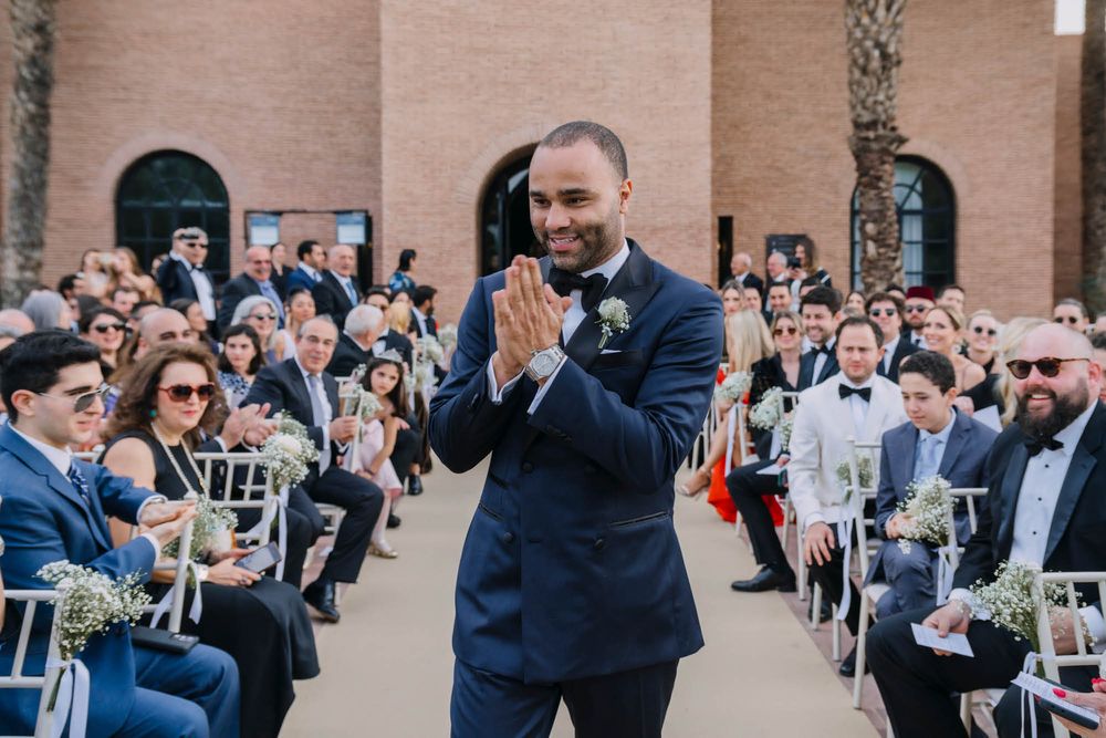 An emotional groom stands at the end of the aisle during an outdoor wedding ceremony with guests seated on both sides.