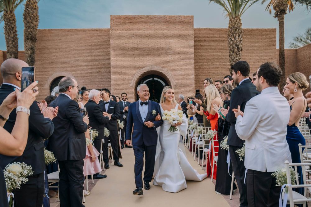 Wedding guests celebrate with cameras and phones as a couple exits a desert venue with reddish clay walls and palm trees.