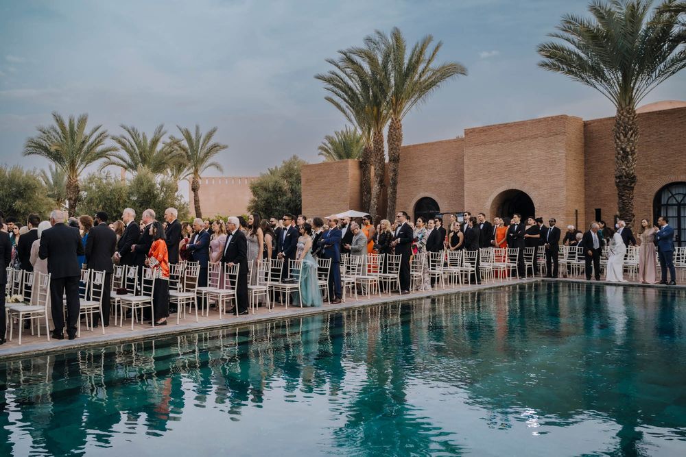 A poolside wedding reception at a desert venue with palm trees and guests gathering around a reflection pool at sunset.