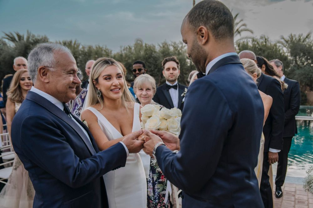 Wedding ceremony guests gather around an outdoor exchange of rings near a swimming pool on a sunny day.