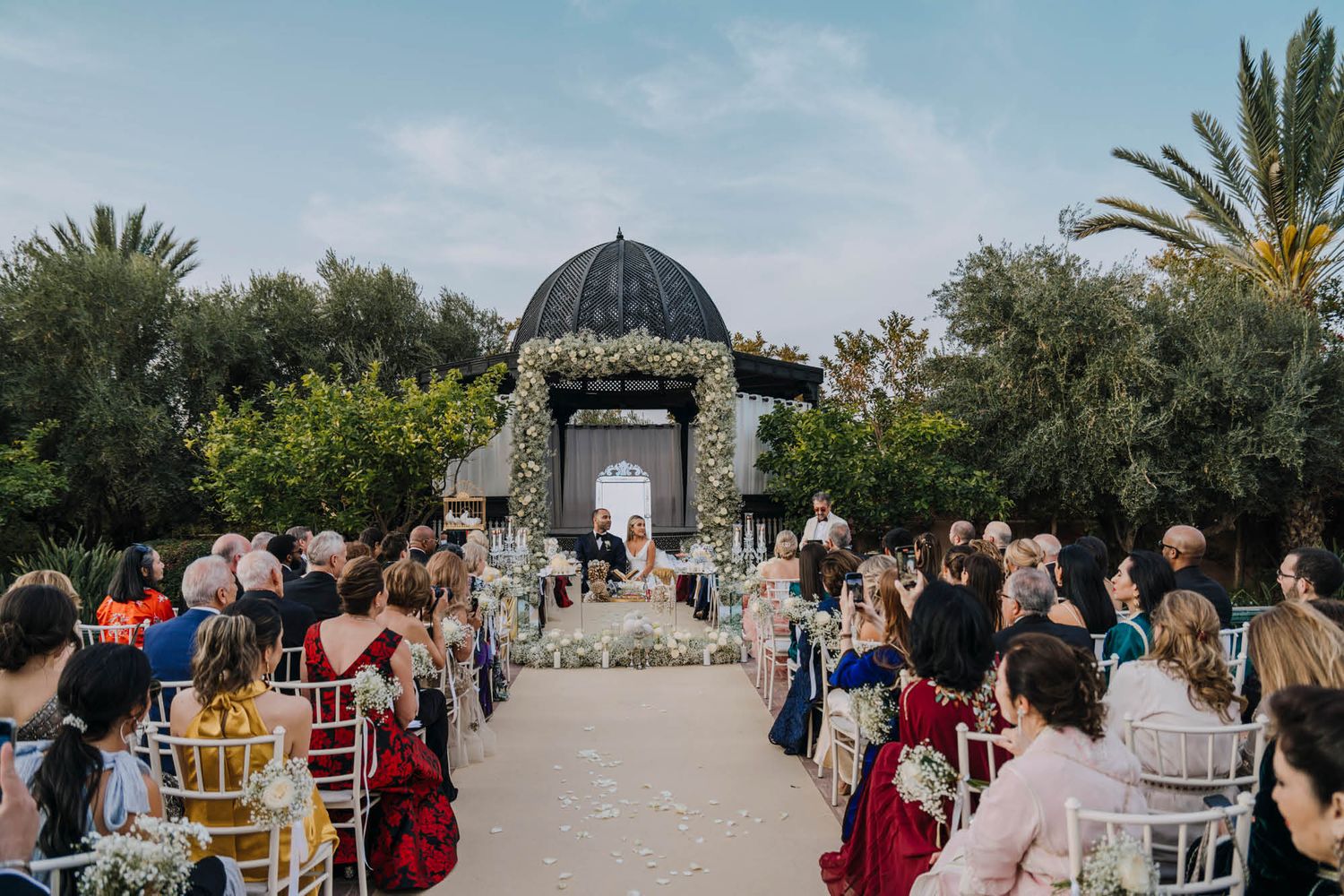 An outdoor wedding ceremony takes place under an ornate domed gazebo with palm trees and guests seated on both sides.