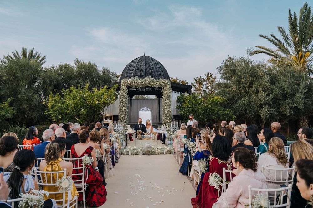 An outdoor wedding ceremony takes place under an ornate domed gazebo with palm trees and guests seated on both sides.