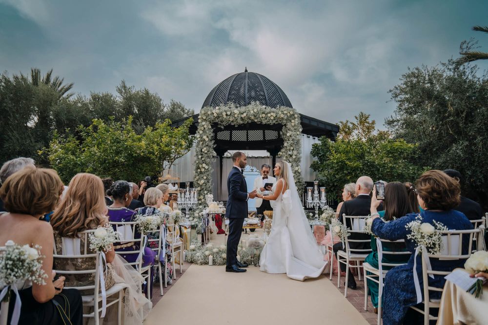 Outdoor wedding ceremony under a domed gazebo with white decor and chairs on a garden pathway.
