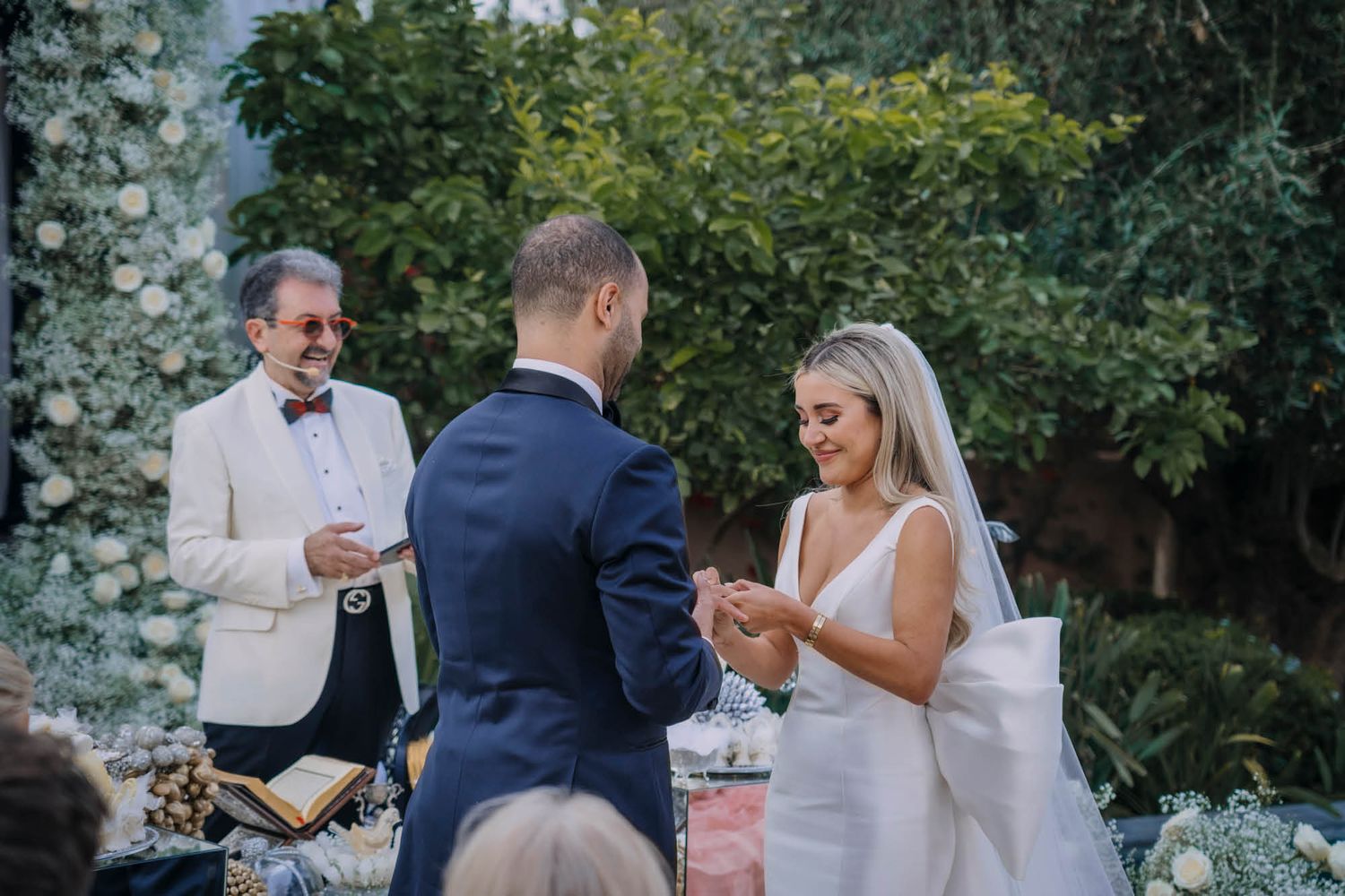 A couple exchanges rings during an outdoor wedding ceremony in a lush garden setting with an officiant present.
