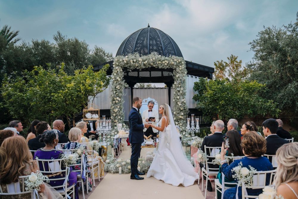An outdoor wedding ceremony takes place under an ornate stone gazebo with guests seated in white chairs along a paved aisle.