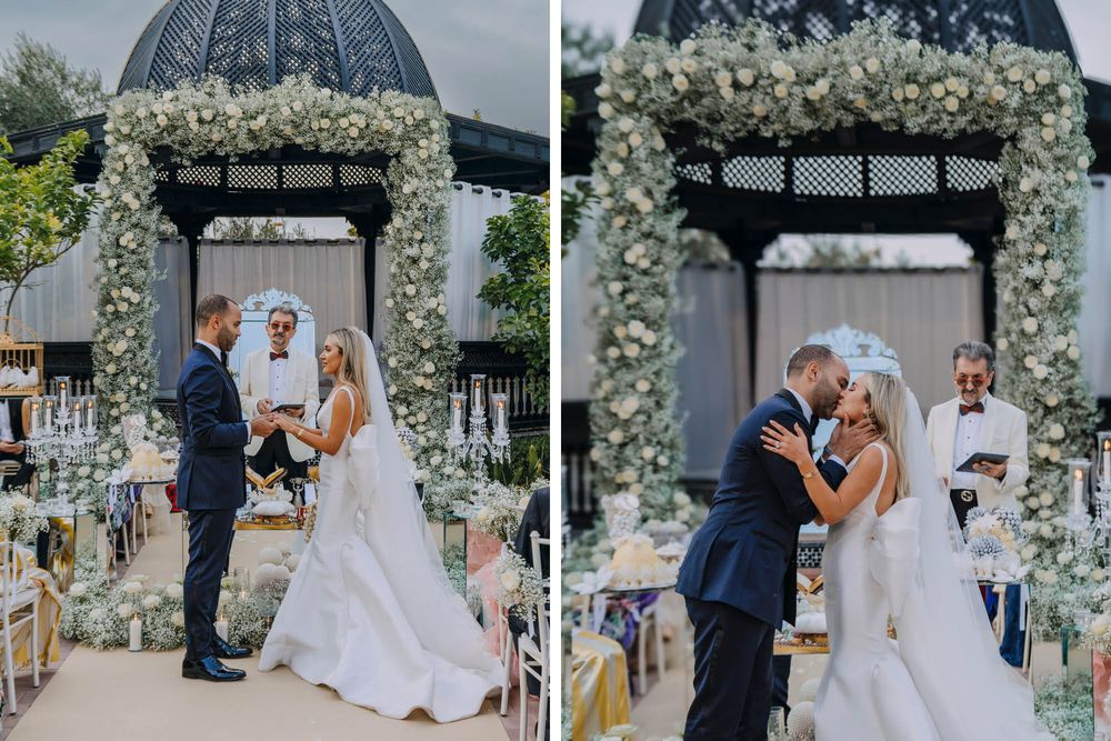 A romantic outdoor wedding ceremony takes place under a domed gazebo decorated with white floral archway decorations.