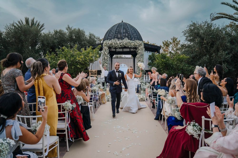 A wedding ceremony takes place under an elegant domed gazebo with guests seated along a garden path.