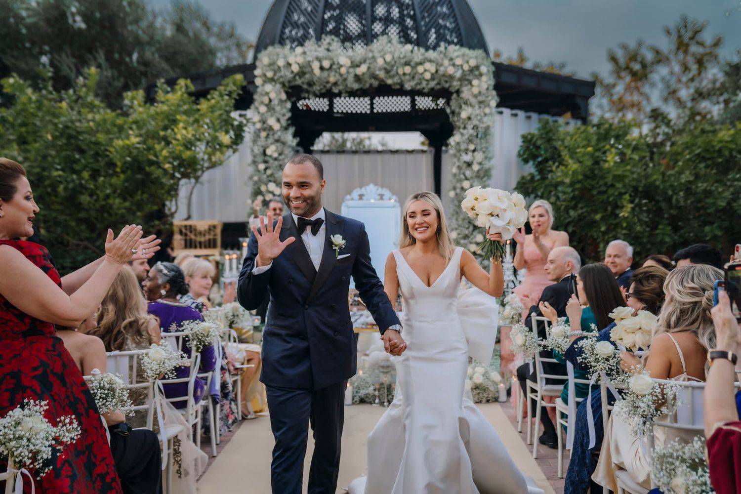 A couple celebrates walking down the aisle at an outdoor wedding ceremony decorated with white floral arrangements.
