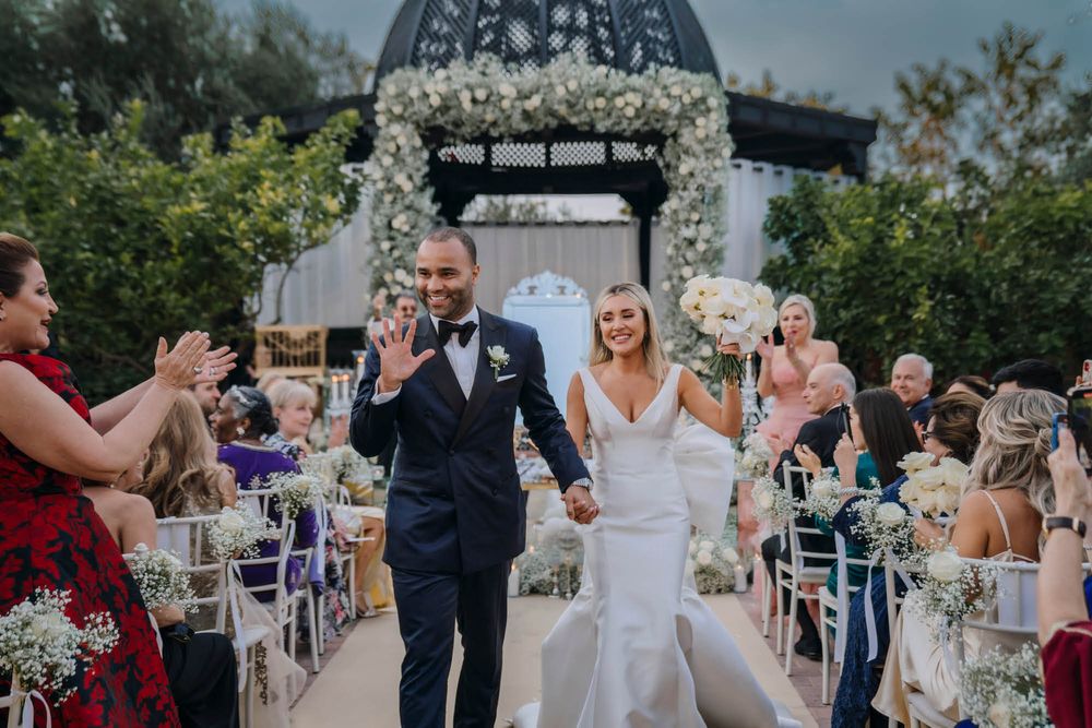 A couple celebrates walking down the aisle at an outdoor wedding ceremony decorated with white floral arrangements.