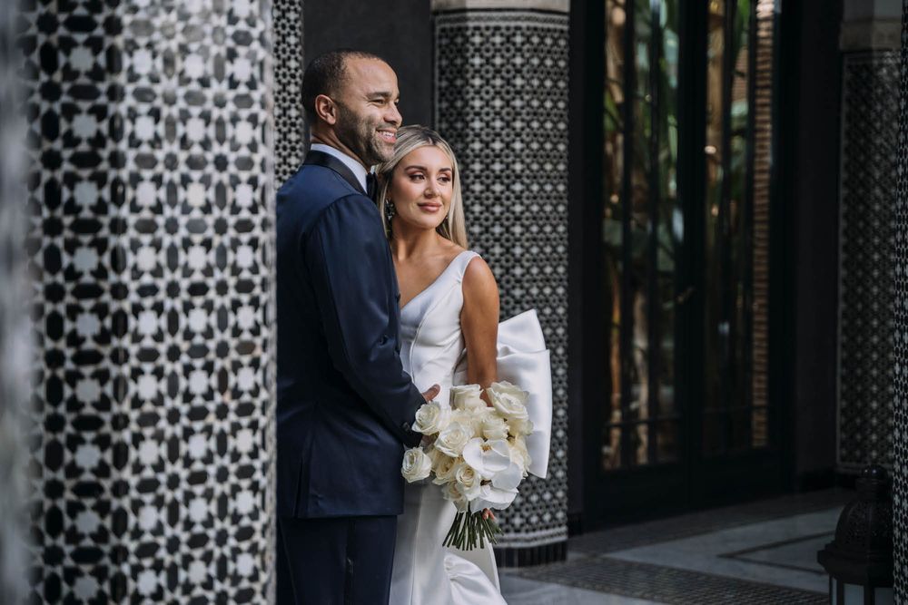 A joyful wedding couple embraces in front of a decorative lattice wall, the bride holding a white bouquet.
