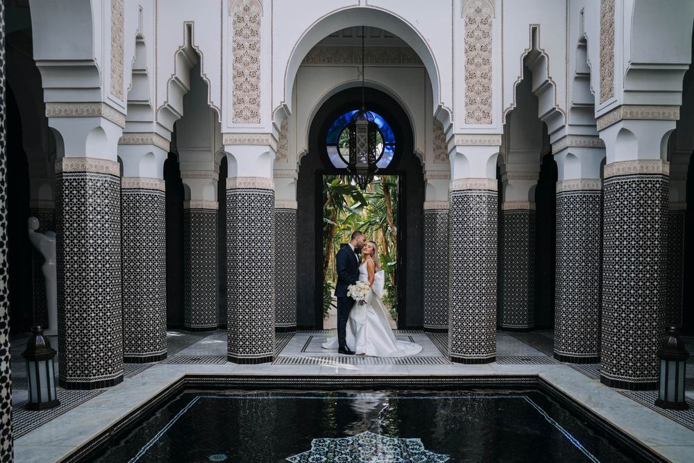 Elegant couple stands beneath ornate Moroccan archways with reflecting pool at luxury La Mamounia Hotel in Marrakech.
