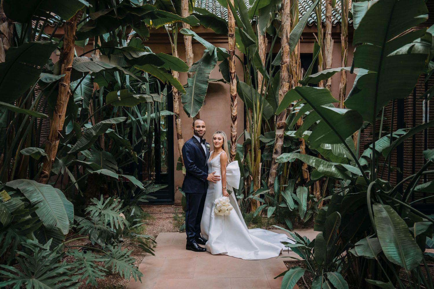 Wedding couple standing on pathway surrounded by lush tropical plants and banana trees.