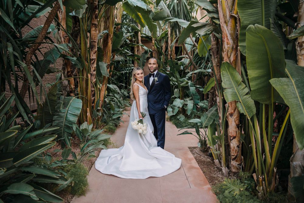 A couple in wedding attire poses along a pathway surrounded by lush tropical plants and banana trees.