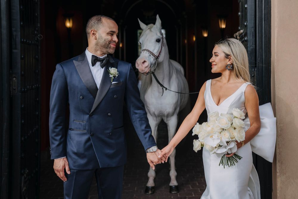 A bride and groom pose with a beautiful white horse in formal wedding attire outside an elegant venue.