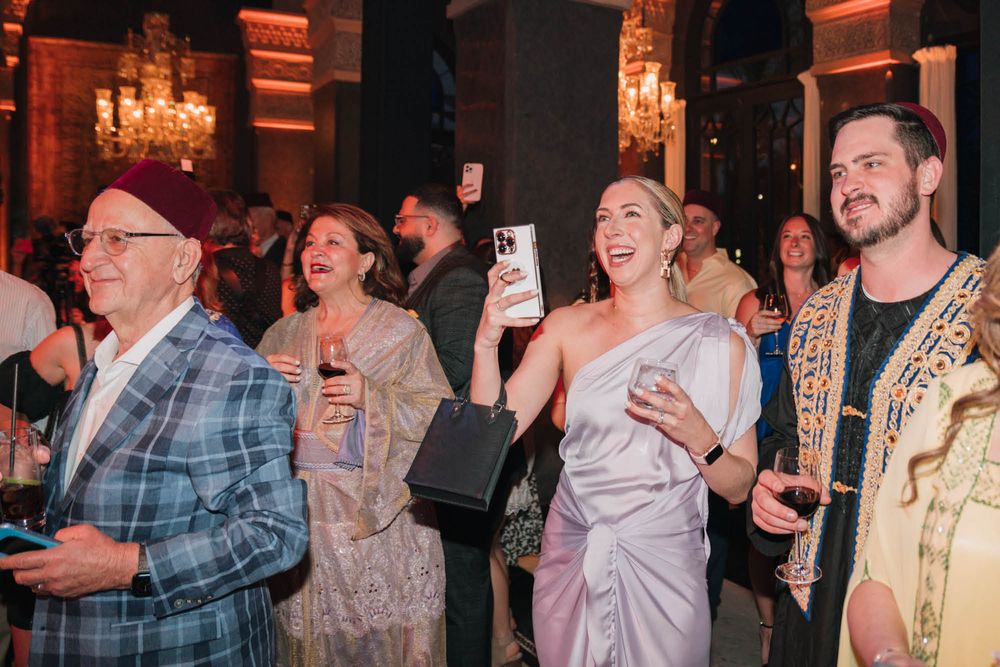 Party guests celebrate and take photos in an elegantly lit ballroom with chandeliers.