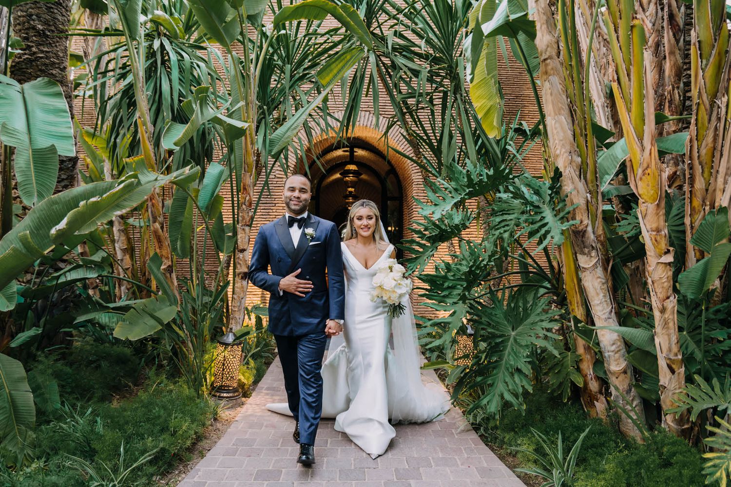 A couple walks down a tropical garden path lined with lush banana trees and palms.